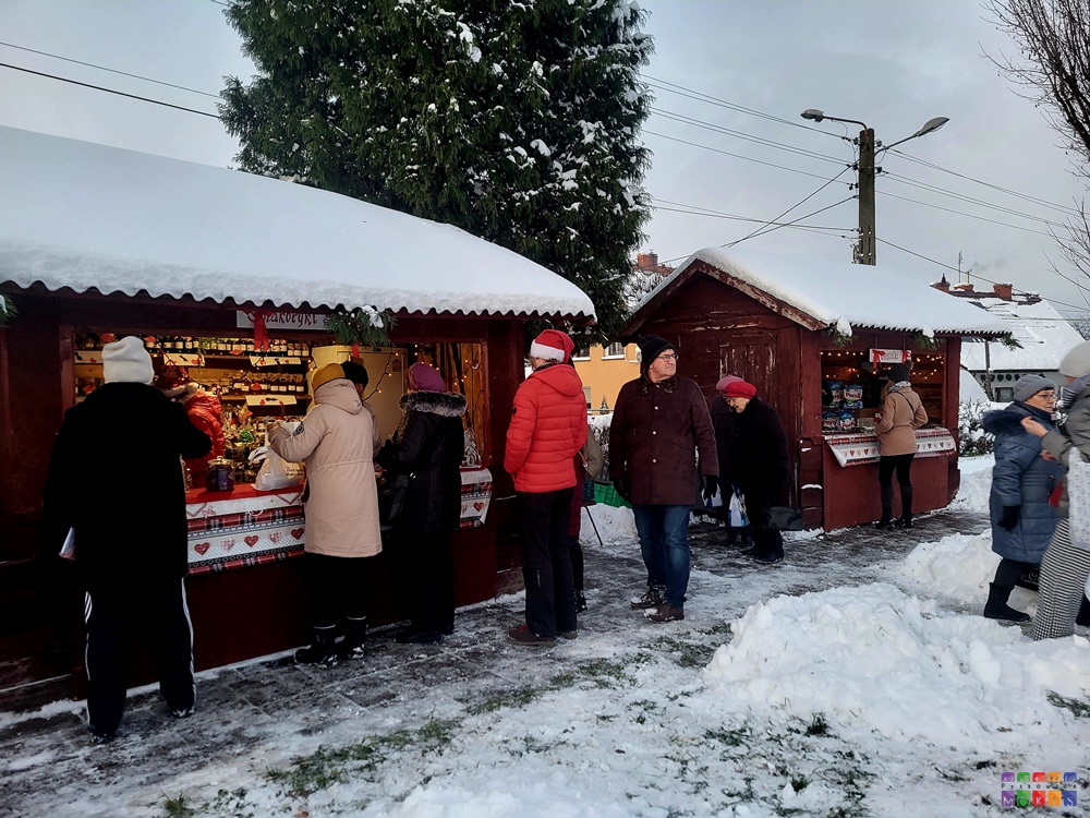 Zdjęcie przedstawiające stragany z ozdobami świątecznymi a przed nimi stojących ludzi i oglądających. Scenografia zimowa.