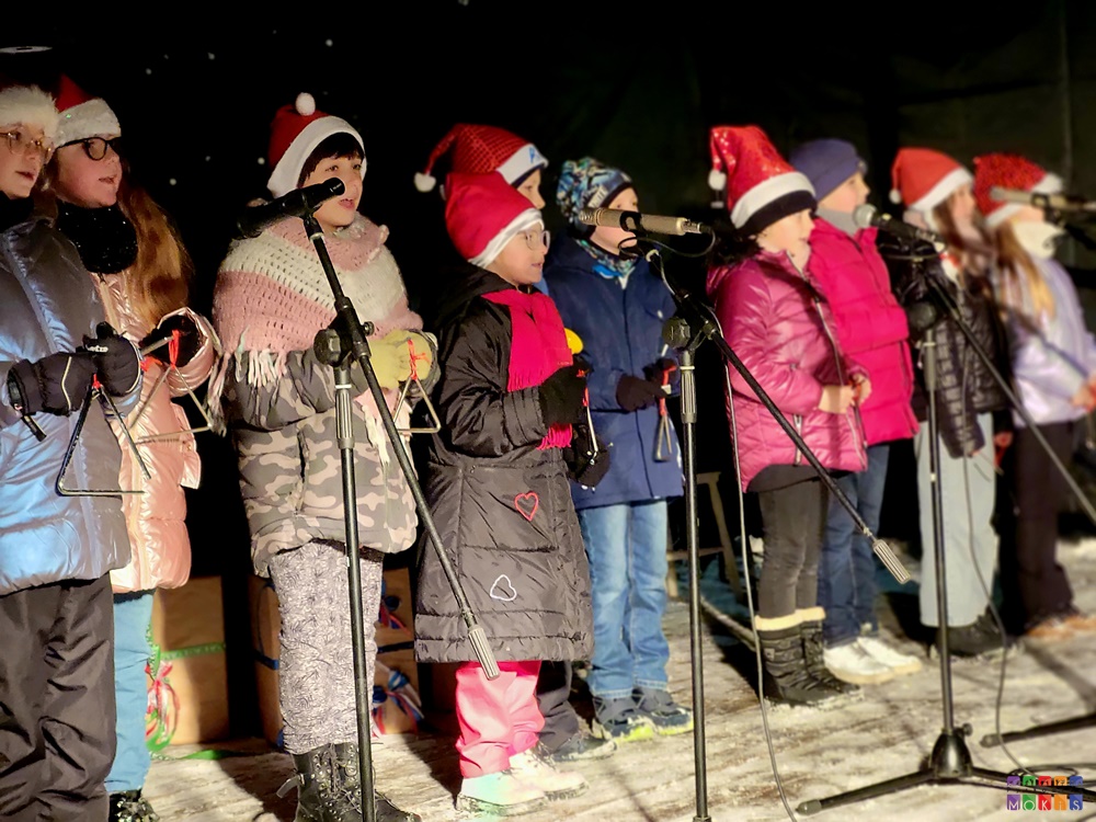 Zdjęcie przedstawiające śpiewające dzieci do mikrofonów. Są one ciepło ubrane.