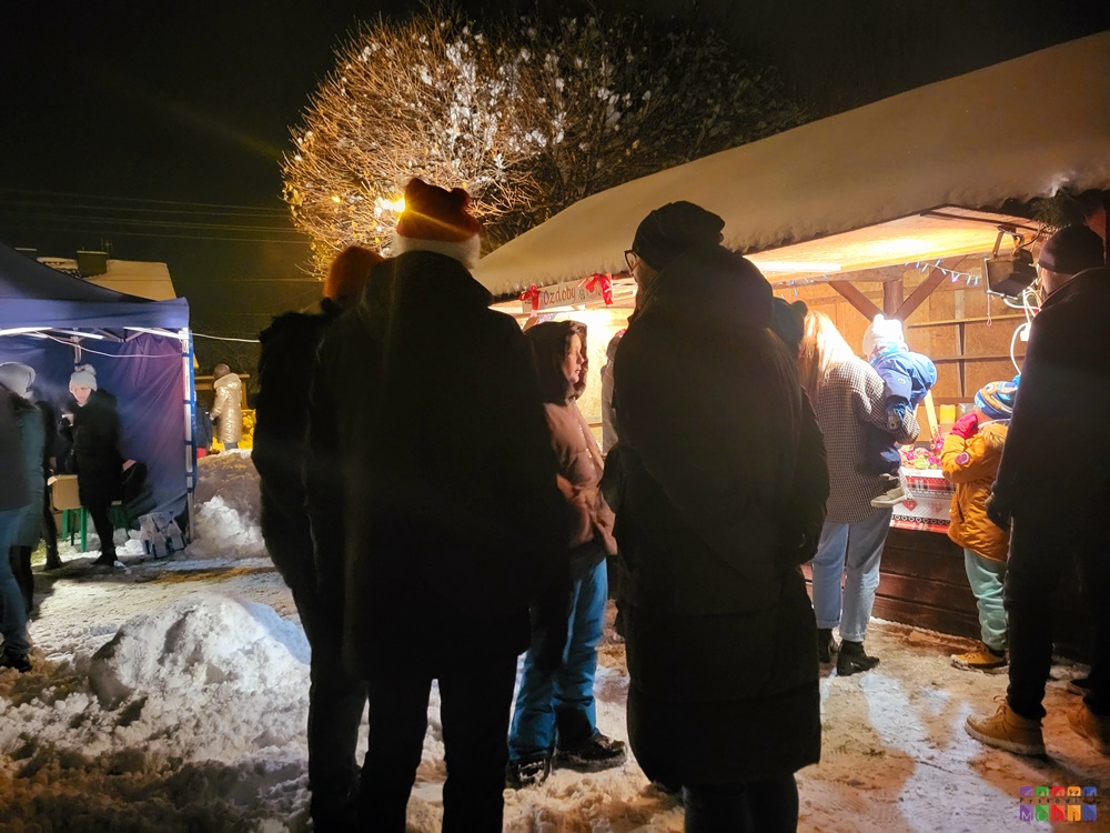 Zdjęcie przedstawiające stragan z ozdobami świątecznymi a przed nim stojących ludzi i oglądających. Scenografia zimowa.
