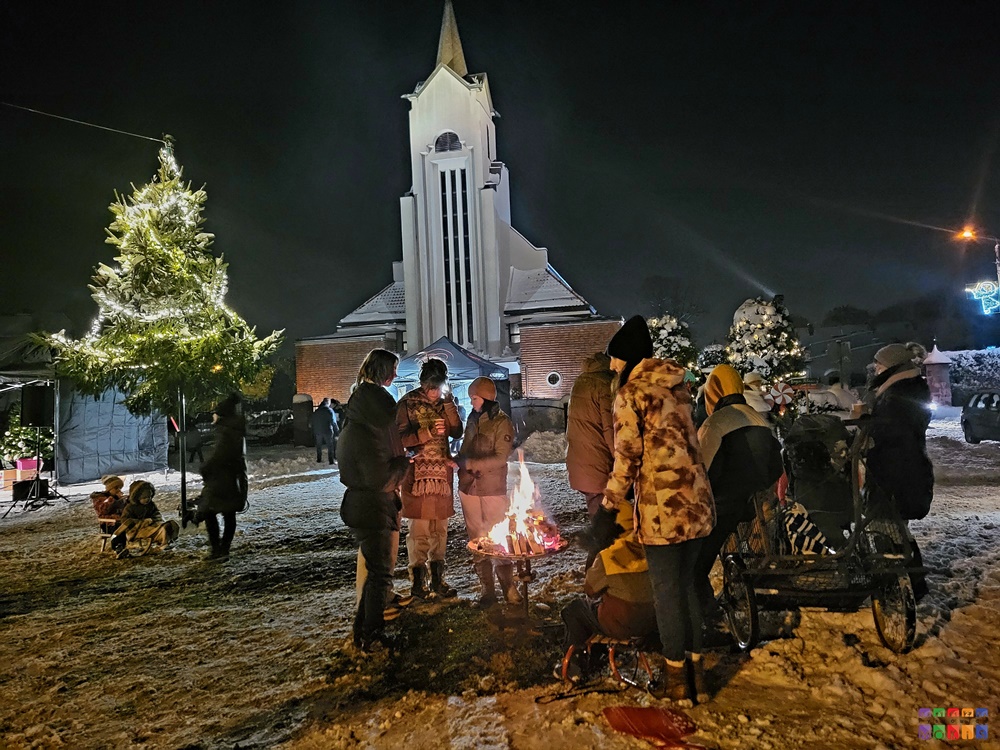 Zdjęcie przedstawia grupę ludzi stojących przy ognisku w scenografii zimowej. W tle widać wieżę kościoła oraz rozświetloną choinkę.