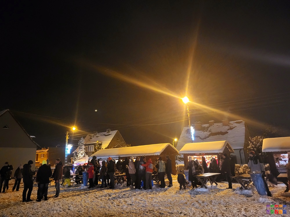 Zdjęcie przedstawiające stragany z ozdobami świątecznymi a przed nimi stojących ludzi i oglądających. Scenografia zimowa.
