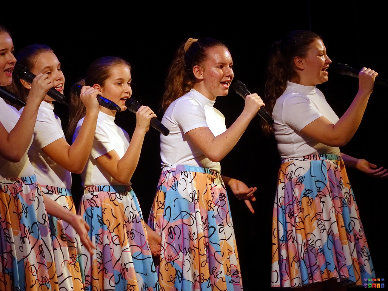 Zdjęcie przedstawia śpiewające do mikrofonów dziewczyny. Mają one białą koszulkę oraz kolorowe sukienki.