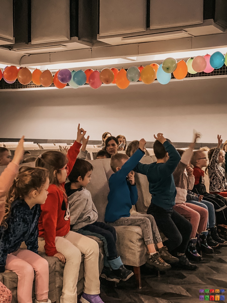 Zdjęcie przedstawiające siedzące i uśmiechające się dzieci w sali widowiskowej MOKiS mają one podniesione ręce nad nimi wiszą balony