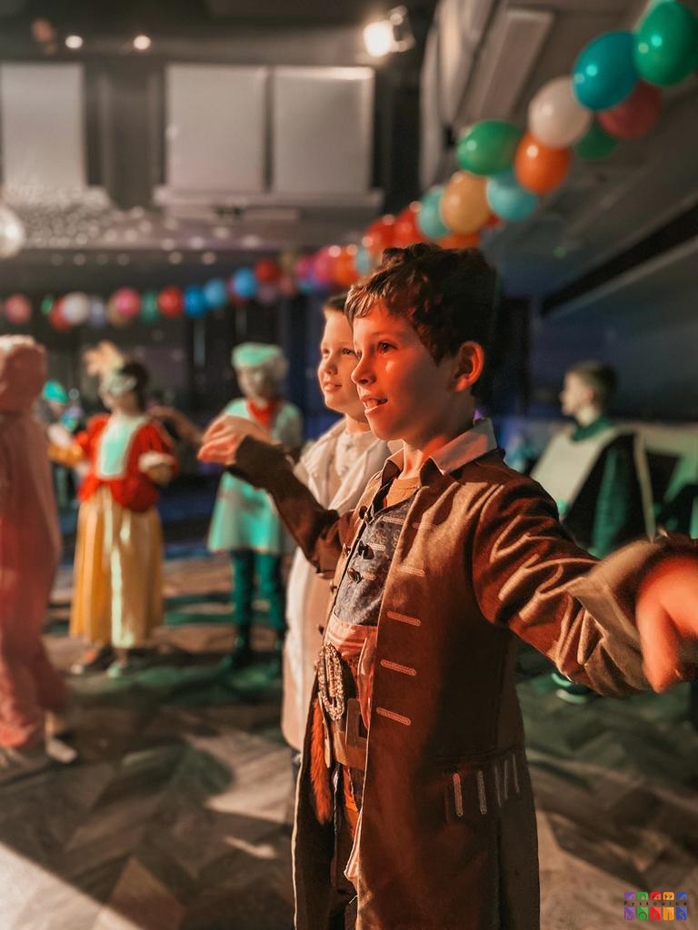 Zdjęcie przedstawiające bawiących się dzieci na balu karnawałowym. Każdy z nich jest przebadany za różne postacie. W koło balony oraz świecące światła.