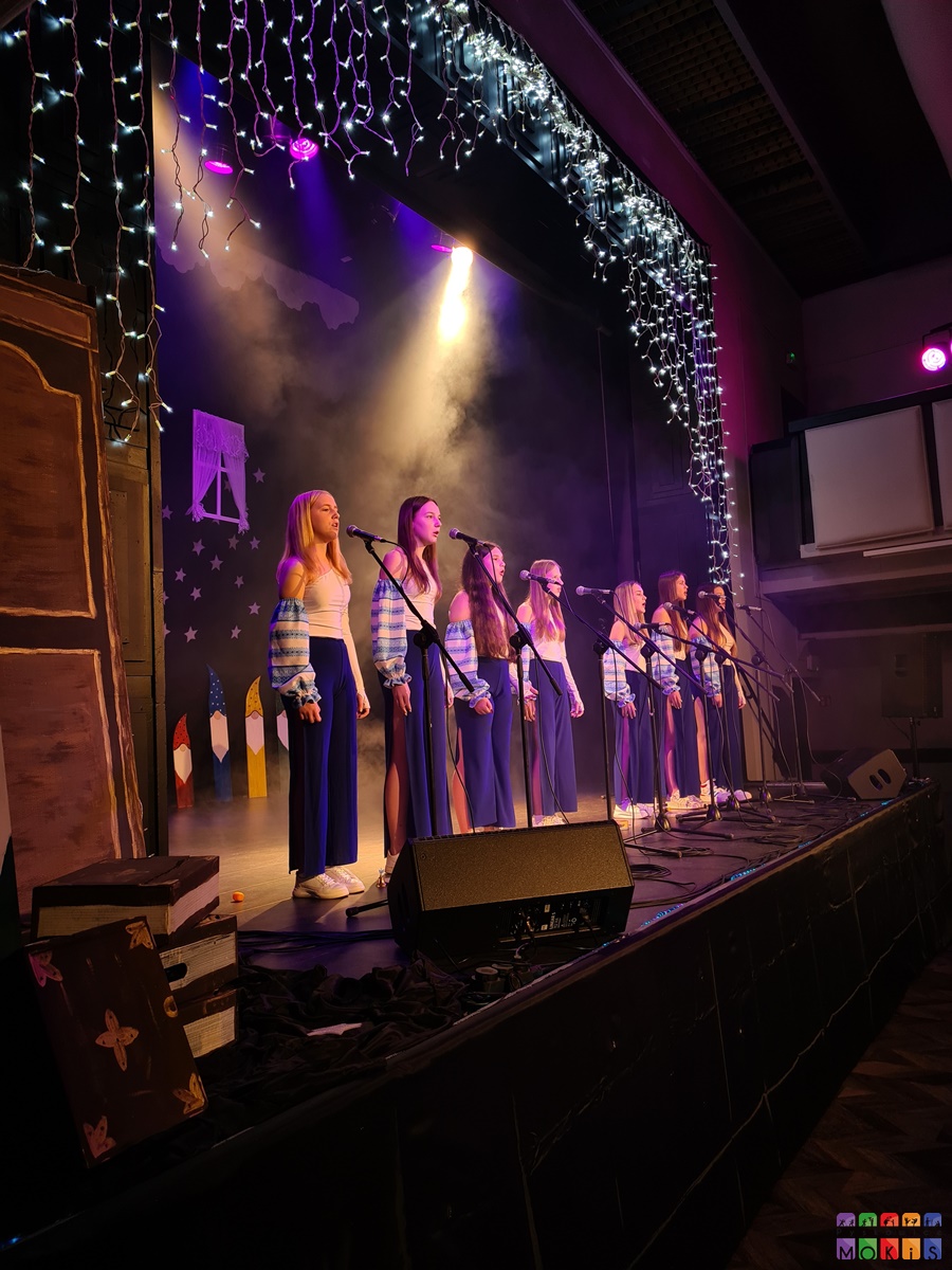 Zdjęcie przedstawia stojących wokalistów na rozświetlonej scenie przy mikrofonach na statywie