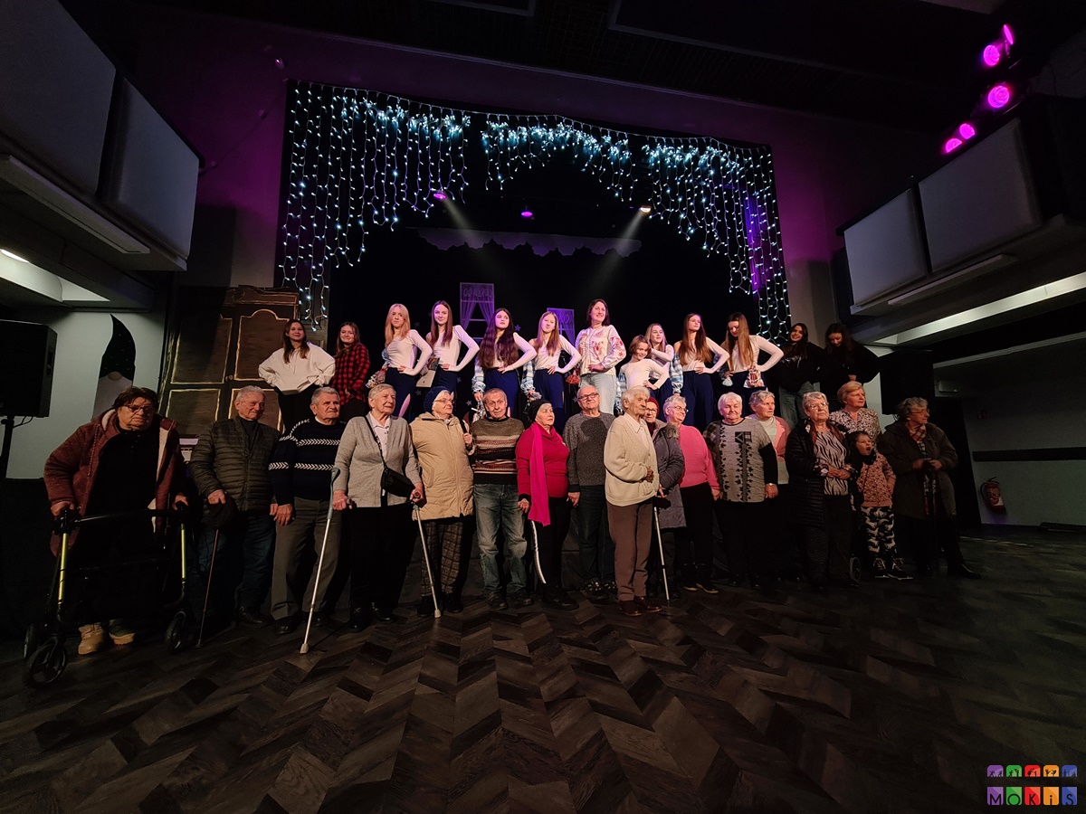 Zdjęcie przedstawia grupowe zdjęcie starszych ludzi z wokalistami na tle rozświetlonej sceny