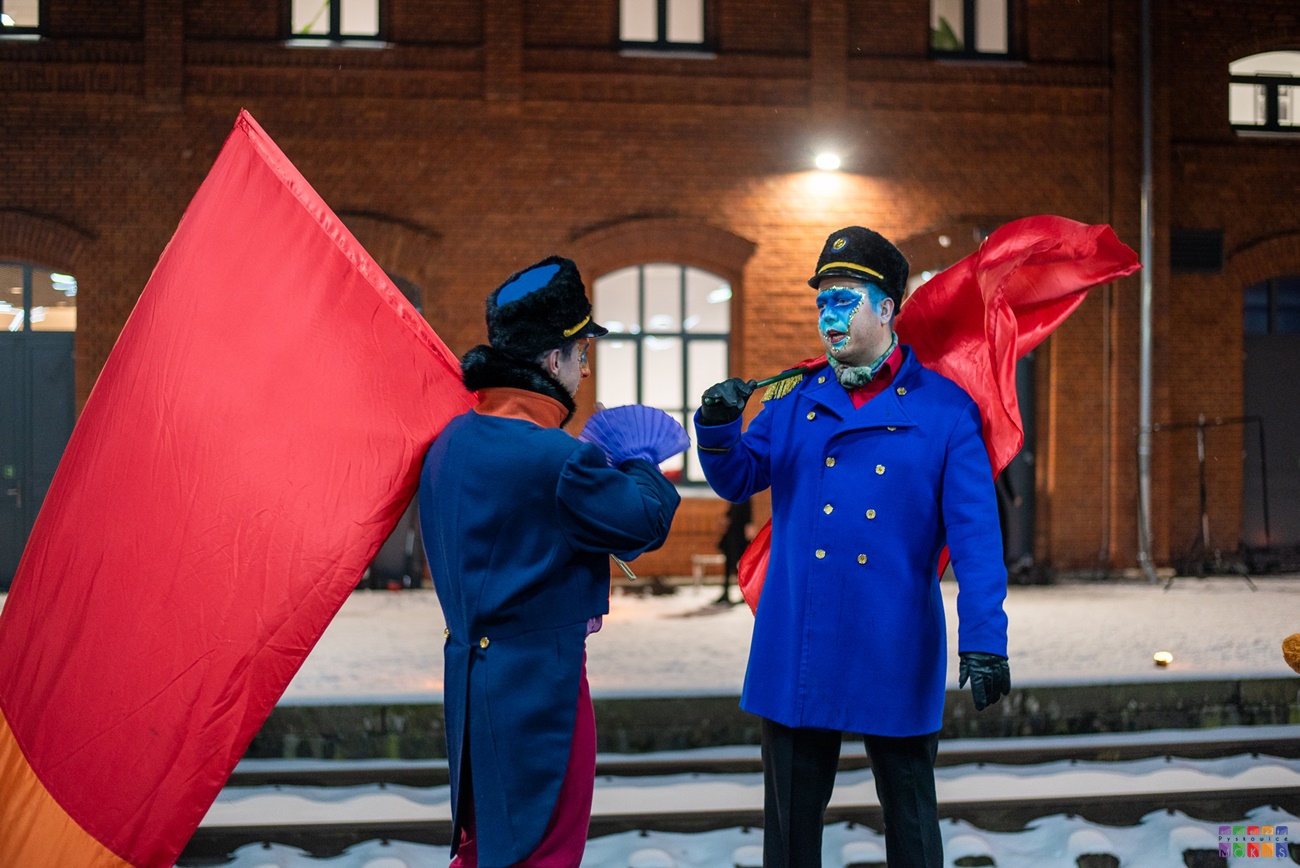 Zdjęcie przedstawia dwóch pomalowanych mężczyzn z czerwonymi flagami na tle budynku Stacji Kultury
