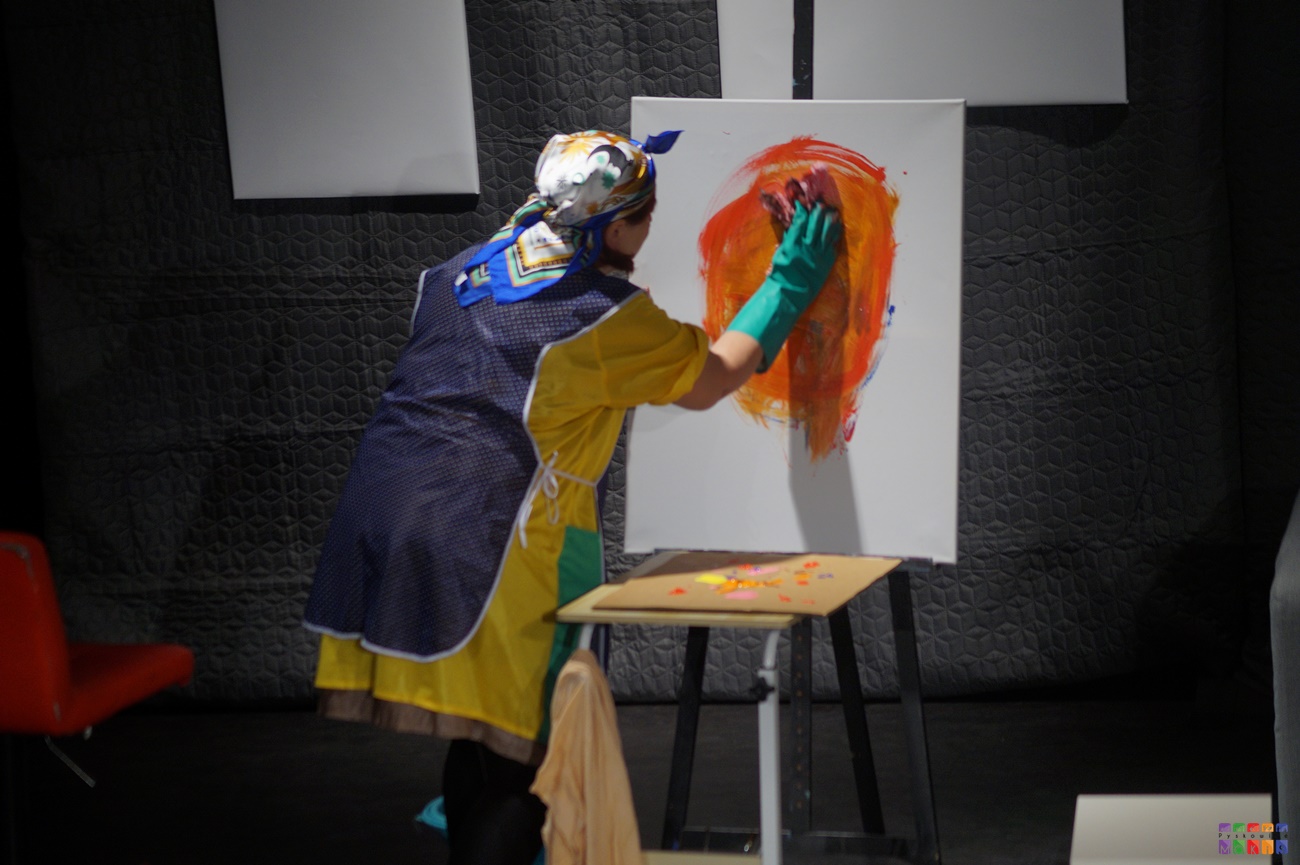 Zdjęcie przedstawia grę aktorską na scenie MOKiS-u. Kobieta przebrana za sprzątaczkę maluje coś na płutnie.
