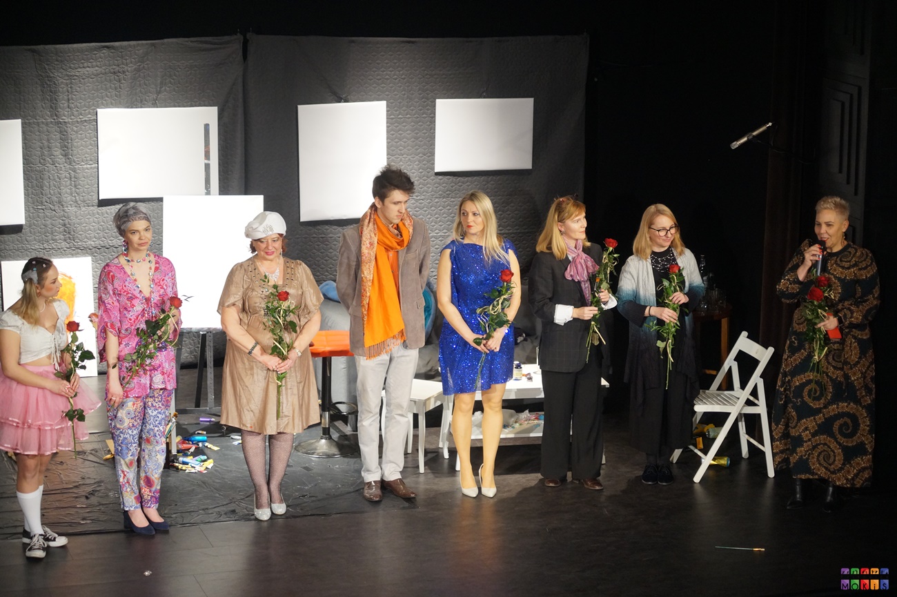 Zdjęcie przedstawia ustawionych w rzędzie aktorów z różami w ręku na tle scenografii do przedstawienia
