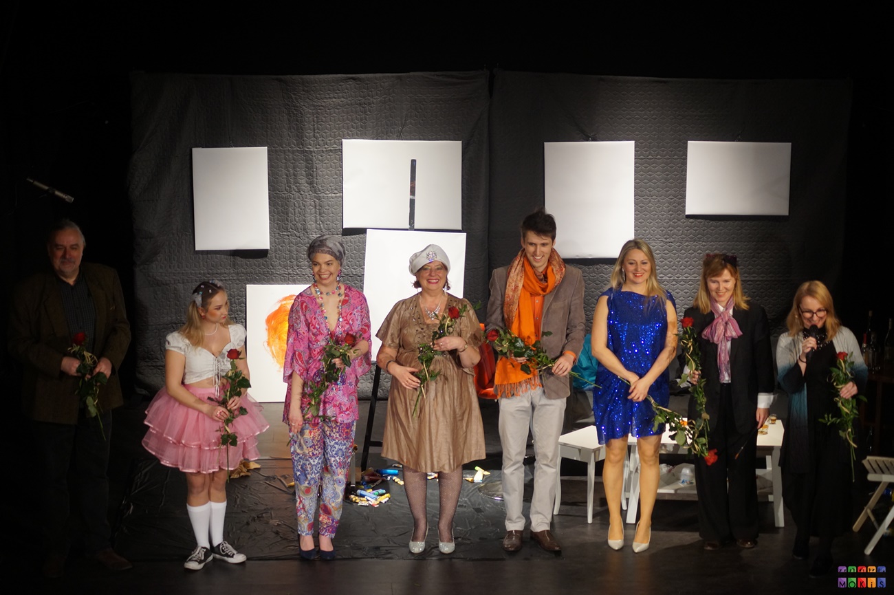 Zdjęcie przedstawia ustawionych w rzędzie aktorów z różami w ręku na tle scenografii do przedstawienia
