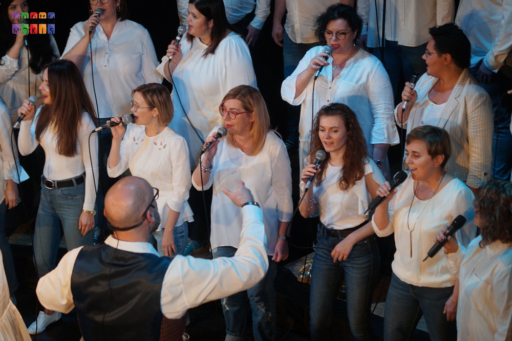 Zdjęcie przedstawia grupę ludzi śpiewających na scenie. Przed nimi obrócony tyłem do fotografa mężczyzna prowadzący zespół