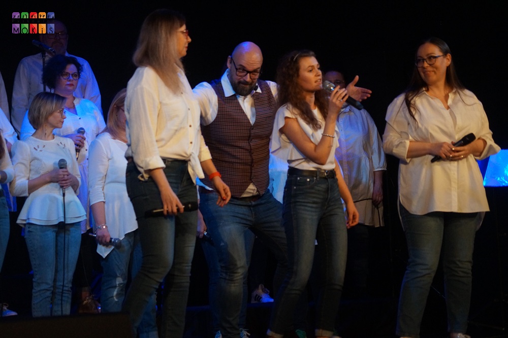 Zdjęcie przedstawia grupę ludzi śpiewających na scenie. Przed nimi trzy wokalistki z mikrofonami