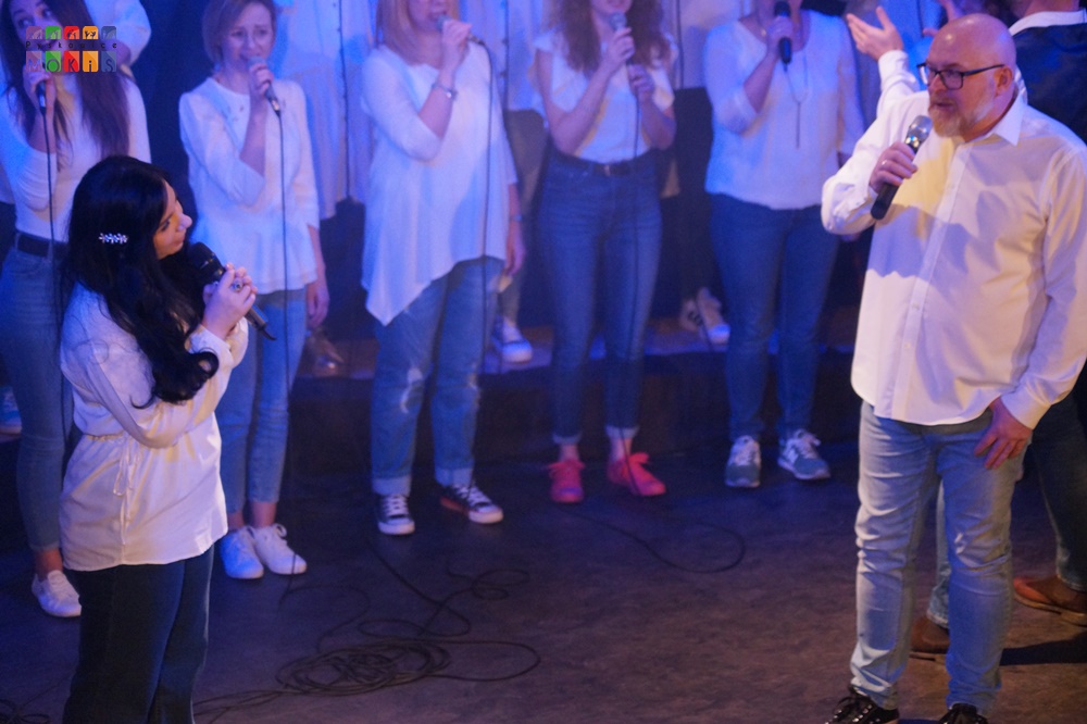 Zdjęcie przedstawia grupę ludzi śpiewających na scenie. Przed nimi dwoje wokalistów mężczyzna oraz kobieta śpiewających do mikrofonu.