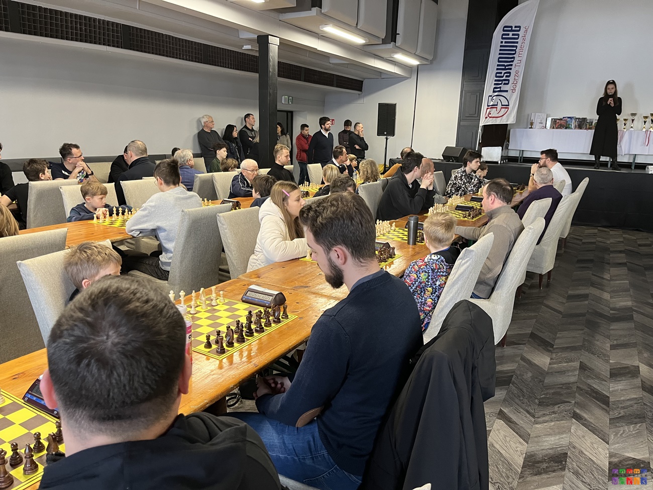 Zdjęcie przedstawia ogólny widok na salę widowiskową gdzie rozstawione są stoły, przy których siedzą ludzie grający w szachy.
