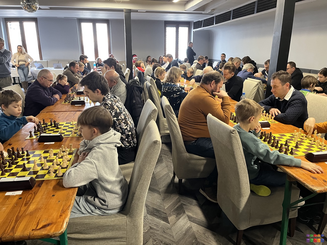 Zdjęcie przedstawia ogólny widok na salę widowiskową gdzie rozstawione są stoły, przy których siedzą ludzie grający w szachy.