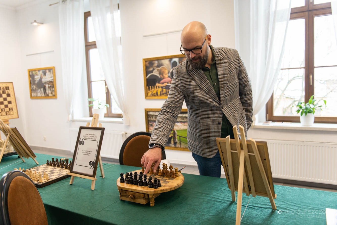 Zdjęcie przedstawia mężczyznę wskazującego na rozłożone szachy palcem.