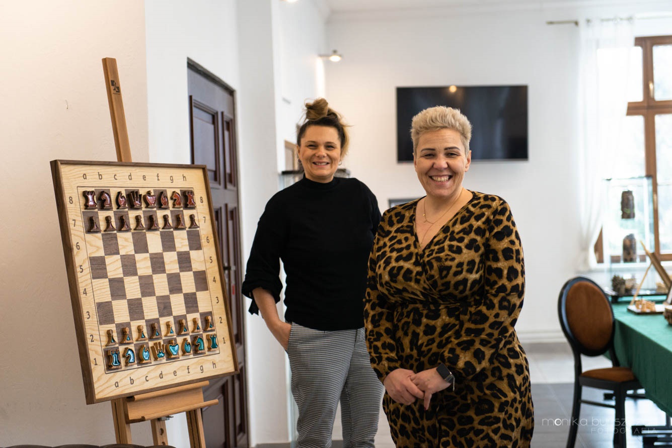 Zdjęcie przedstawia dwie uśmiechnięte kobiety. W tle wystawa szachów.