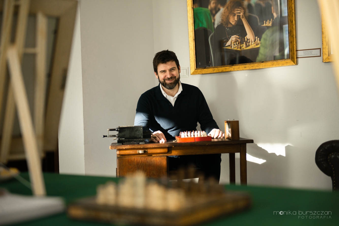 Zdjęcie przedstawia mężczyznę siedzącego przy stoliku z rozłożonymi szachami. Uśmiecha się do fotografa