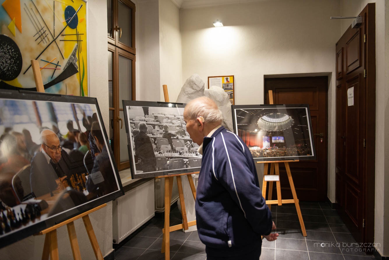 Zdjęcie przedstawia starszego mężczyznę oglądającego wystawę obrazów o tematyce szachowej.